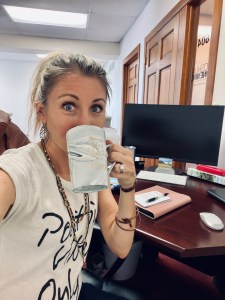 Jessica looking wide eyed and excited behind a coffee mug that says "Let the Adventure Begin". She is sitting at a desk at her new job.