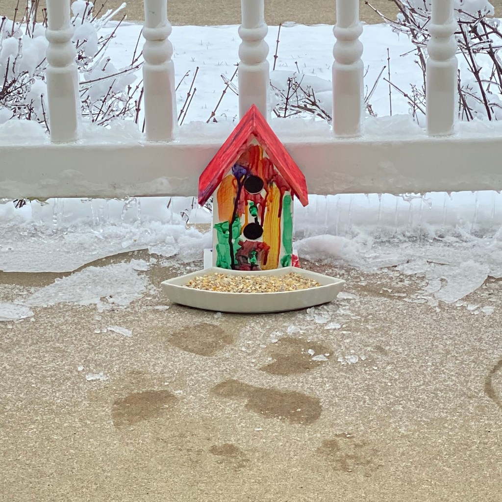 A small birdhouse painted in a variety of colors with two holes, sitting on a porch in the snow with a bowl of birdseed in front.