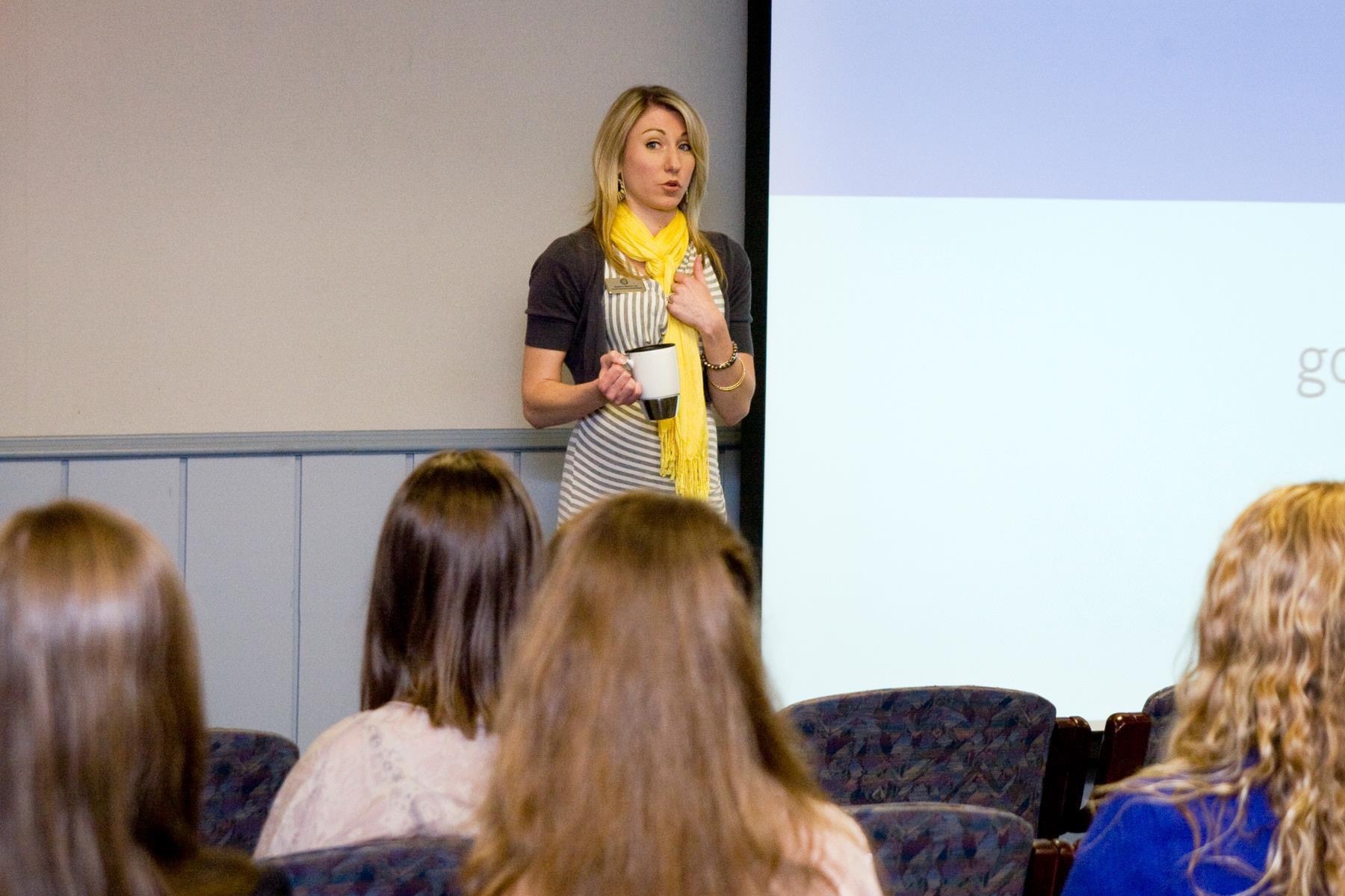 Blonde woman in a yellow scarf giving a standing presentation in front of a screen with a coffee mug in her hand