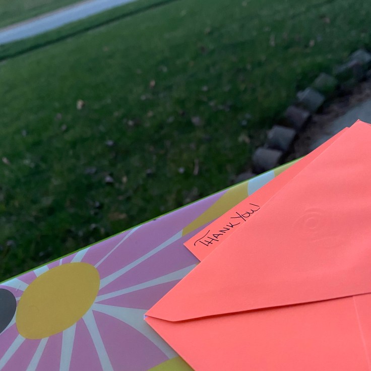 neon orange notecard with the handwritten words Thank You in the foreground, grassy yard in the background